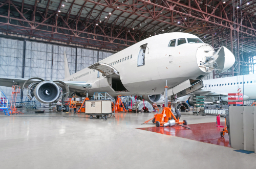 A large commercial aeroplane in a spacious hangar undergoing maintenance, with its doors and engine panels open and various equipment and scaffolding placed around it.