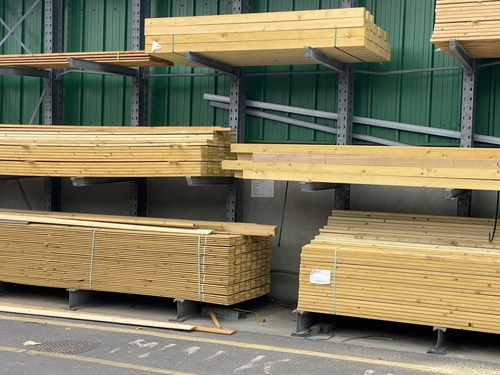 Stacks of wooden planks are neatly arranged on metal racks against a green wall at a timber yard or hardware shop. Some planks are bound with straps, and a few loose pieces are on the ground.