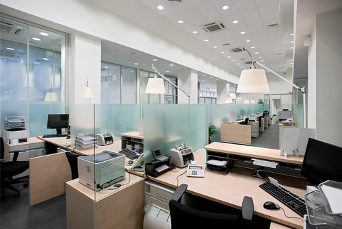 Modern open-plan office with rows of desks separated by frosted glass partitions, computers, telephones, printers, and office equipment on each desk. Bright lighting and large windows create a clean, organised workspace.