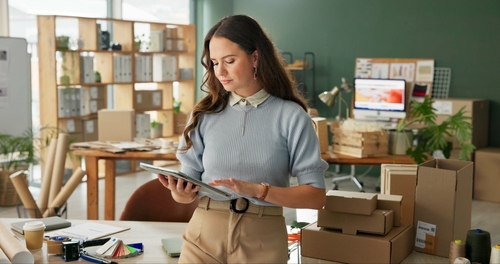 A woman stands in an office filled with boxes and shelves, looking at a digital tablet. The workspace appears organised, with packing materials, a computer, and coffee cup visible on the desk.