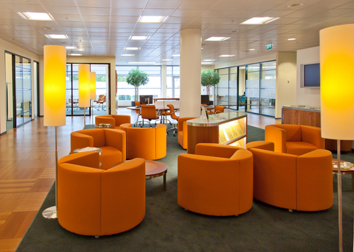 Modern office lounge with bright orange chairs arranged in circles, tall glowing floor lamps, wooden flooring, large windows, and indoor potted trees in the background. The space looks clean and inviting.