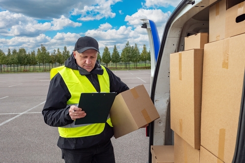 A delivery worker in a yellow safety vest and cap holds a clipboard and a cardboard box, standing next to an open van filled with boxes in a car park with trees and a cloudy sky in the background.