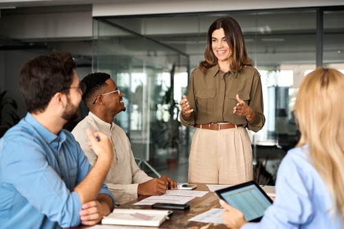 A woman stands and speaks confidently to three colleagues seated around a table in a modern office, whilst they listen and engage. One holds a tablet, another has a notebook, and papers are spread on the table.