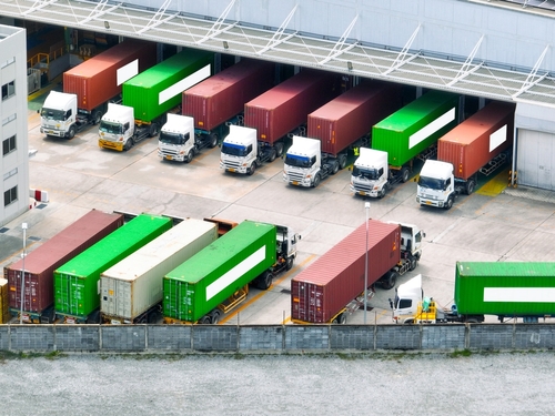 Aerial view of several lorries with red and green shipping containers parked at a loading bay outside a large warehouse building.