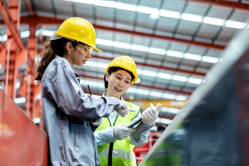 Two women in safety gear, including yellow hard hats and reflective vests, discuss work whilst looking at a tablet in an industrial factory setting.