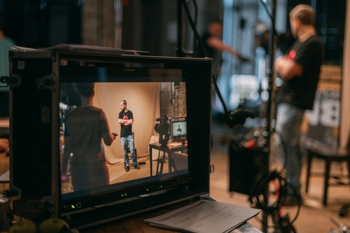 A video monitor displays a man standing and talking in a studio, whilst the man and equipment are visible in the blurred background. Papers and cables are scattered on the studio floor.