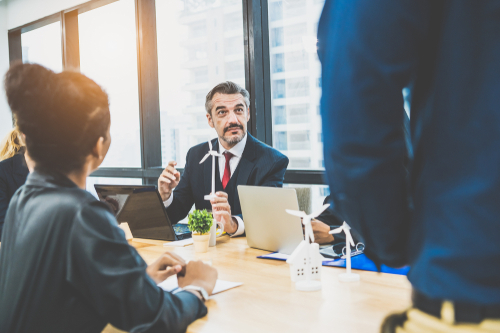 A group of business professionals sits around a table in a modern office, discussing renewable energy. A man in a suit holds a small wind turbine model whilst others listen and take notes.