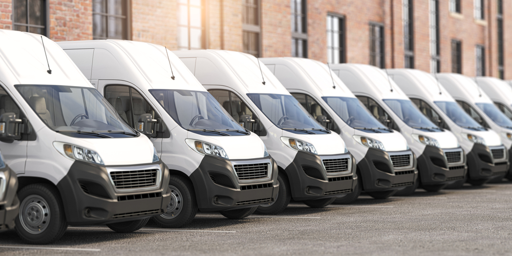 A row of white delivery vans parked side by side in a car park, in front of a brick building. The vans are identical and neatly aligned, facing the same direction.