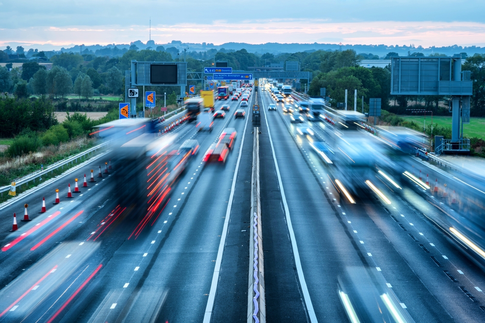Blurred cars travel quickly along a busy multi-lane motorway at dusk, with traffic in both directions and green fields and trees lining the road in the background.