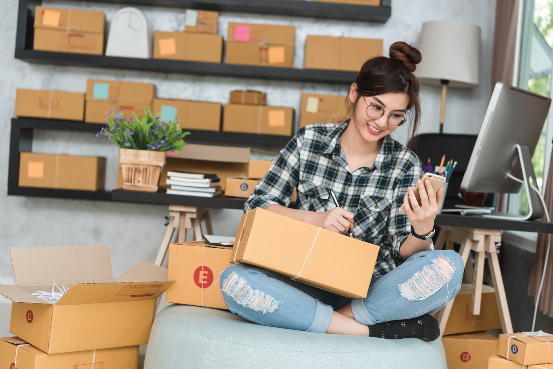 A young woman wearing glasses and ripped jeans sits cross-legged, holding a pen and a cardboard box, looking at her mobile. She is surrounded by parcels in a home office with shelves of boxes behind her.